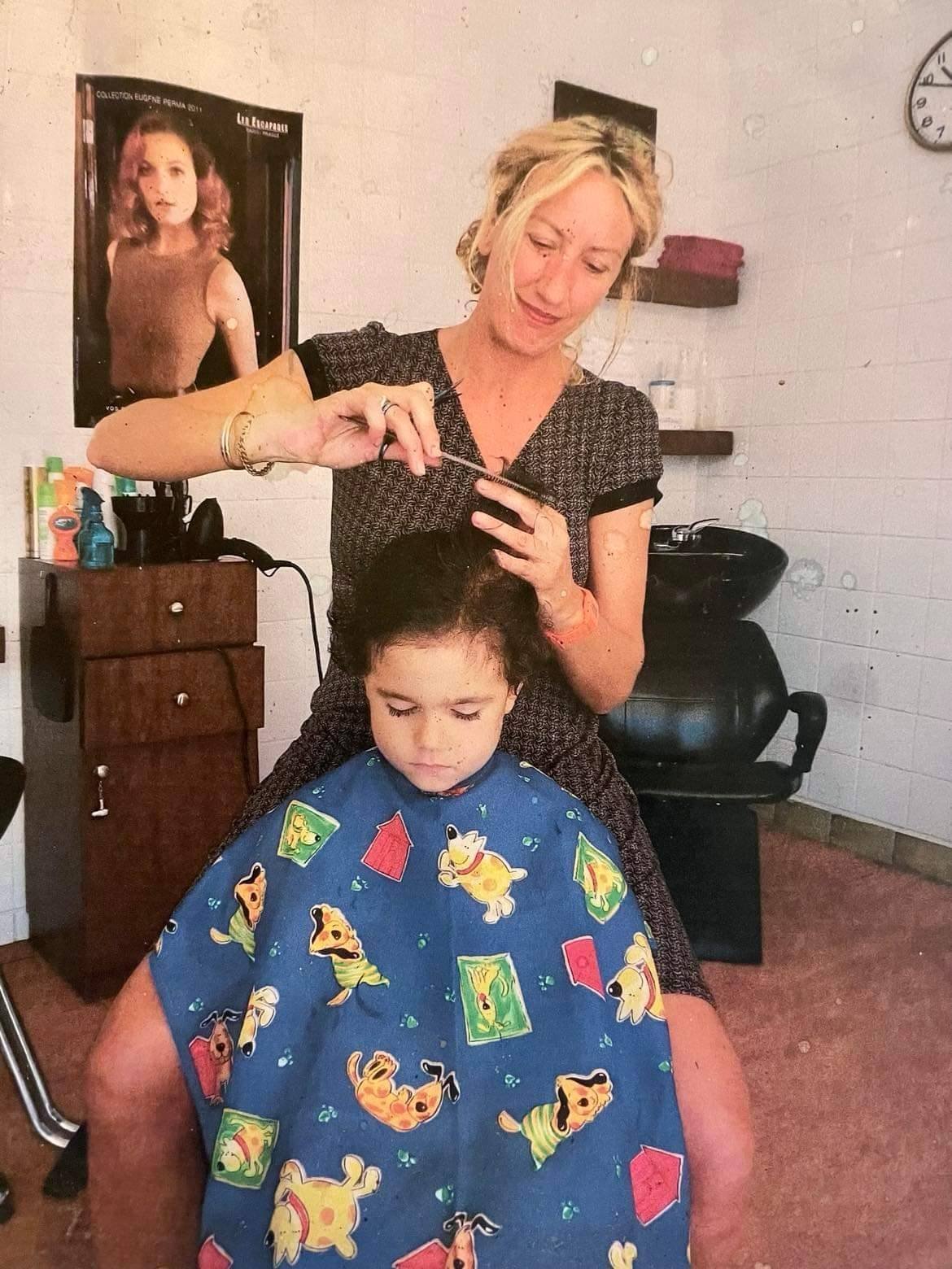 Une coiffeuse coupe les cheveux d'un enfant chez Coiffure Mile-End, affiche vintage au mur.